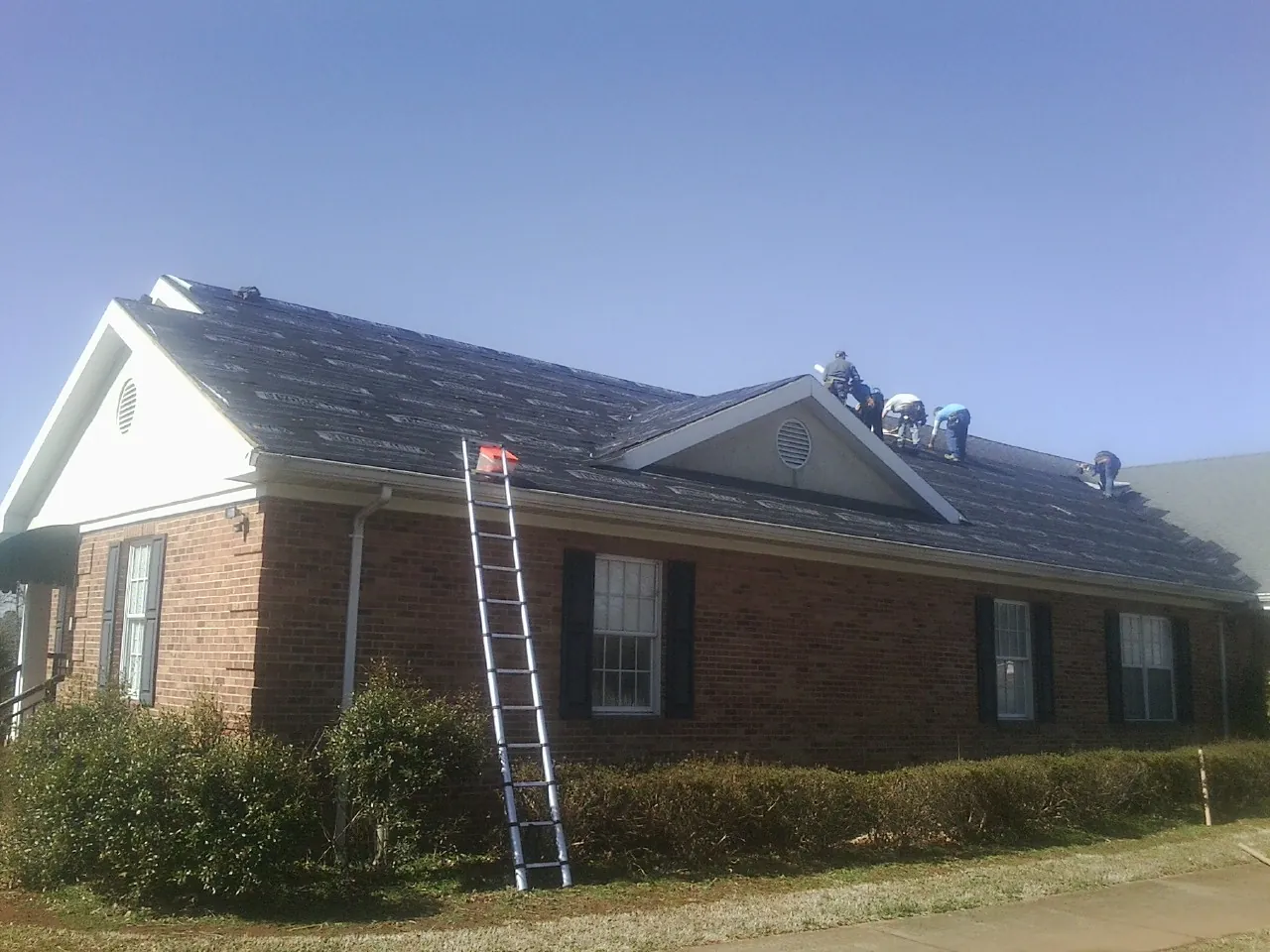 Crew tearing shingles off a brick ranch roof with a ladder set — Metal Roof Repair work by our crew in Clifton